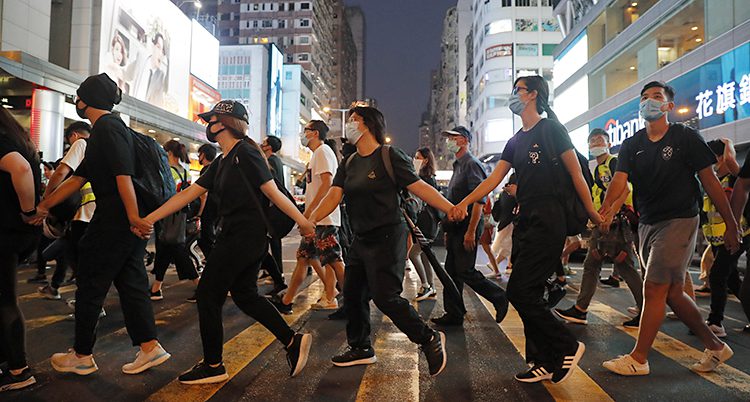 Vi ser en bild från Hongkong. Folk går över ett övergångsställe. De håller varandra i händerna och har masker för ansiktet.