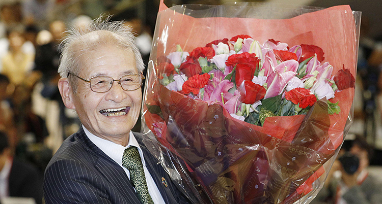 En äldre japansk man som ler. Han har glasögon, kostym och slips. I sin hand håller han en stor blombukett som han har fått.