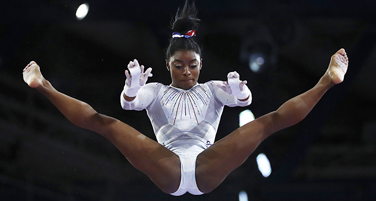 Simone Biles flyger i luften. Hon har båda benen utsträckta. Hon har en vita gymnastikdräkt och en svart knut i håret.