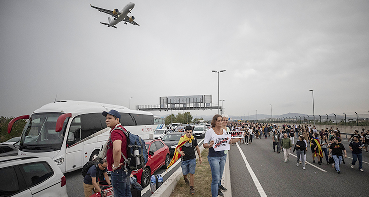 Bilden är tagen nära Barcelonas flygplats. En massa folk går på ena sidan av vägen. Den är blockerad. Inga bilar kan köra där. På andra sidan vägen kör bilar. Ovanför i luften syns ett flygplan.