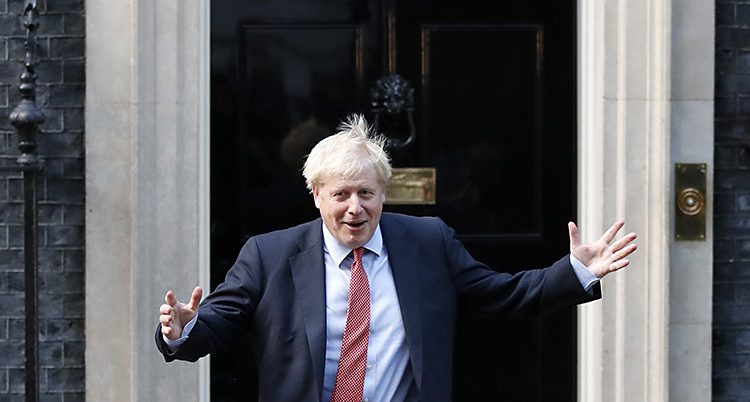 Boris Johnson ser glad ut. Han har båda händerna i luften. Han står framför en dörr.