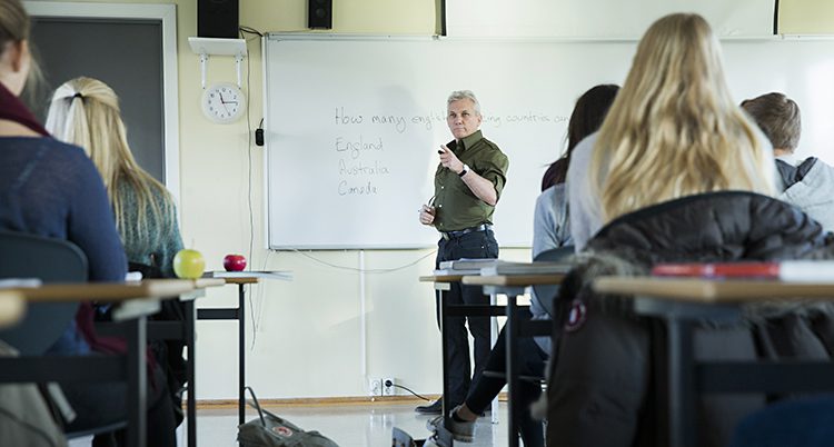 En lärare står vid en tavla i ett klassrum. Några skolbänkar syns.