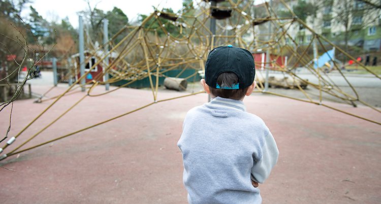 Ett barn står ensam i en lekpark. Bara ryggen syns på barnet.