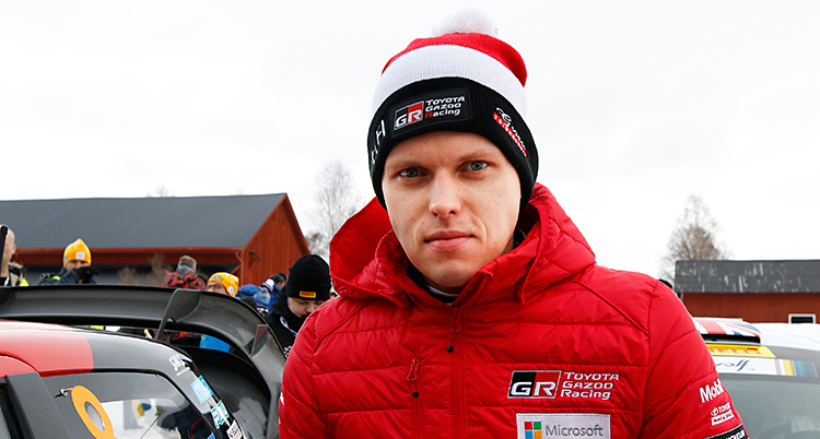 Ott Tänak står bredvid sin rallybil. Han har på sig en tjock röd jacka. På huvudet har han en mössa som är randig i svart, vitt och rött. Mössan har en vit tofs längst upp.