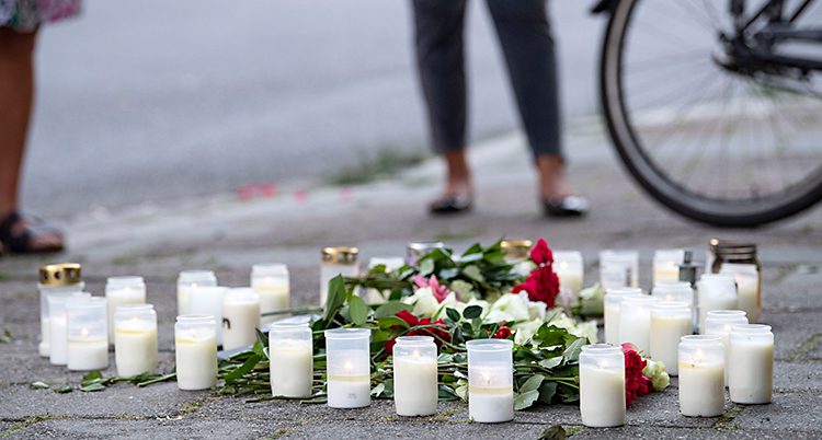 Bilden visar en trottoar. Där finns det blommor och flera tända ljus. Två personer och en cykel står lite längre bak. Man ser bara benen på personerna.