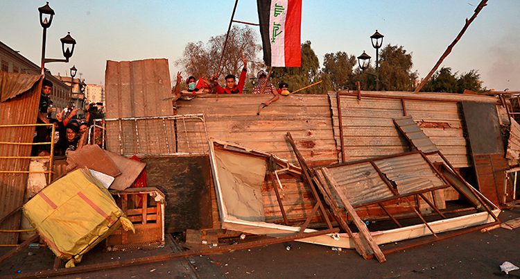 Bilden visar en väg i Bagdad. Folk som protesterar har ställt upp en blockad av trä och annat. Bakom blockaden står demonstranter. De har också en irakisk flagga.