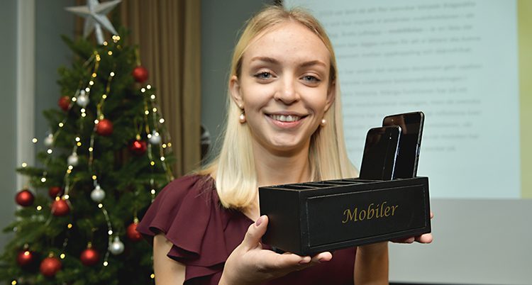 En kvinna framför en julgran håller upp en svart låda med fack där det sticker upp en mobil.