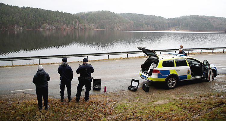 Poliser står vid en väg. Bredvid vägen syns en siö och en polisbil. De styr en drönare.