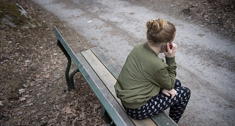 Ryggen på ett barn som sitter på en parkbänk o pratar i telefon.