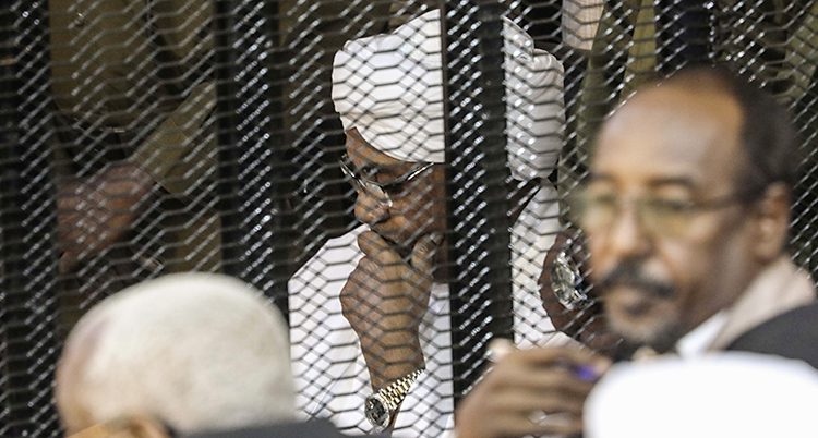 Bilden är från en domstol. Det är en rättegång mot Omar al-Bashir som var ledare i Sudan. Han sitter i en bur. Han har vita kläder och en vit turban på huvudet och glasögon.