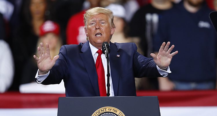 Donald Trump håller ett tal på ett stort möte för folk som gillar honom och hans parti. Han står vid ett podium och pratar i en mikrofon. Han håller ut båda händerna i en gest. Han har på sig en mörkblå kostym och en röd slips.