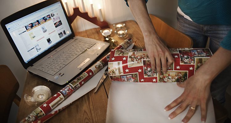 Någon slår in en julklapp i rött papper. Vid sidan om står en uppslagen dator. En webbsida för handel syns på skärmen.