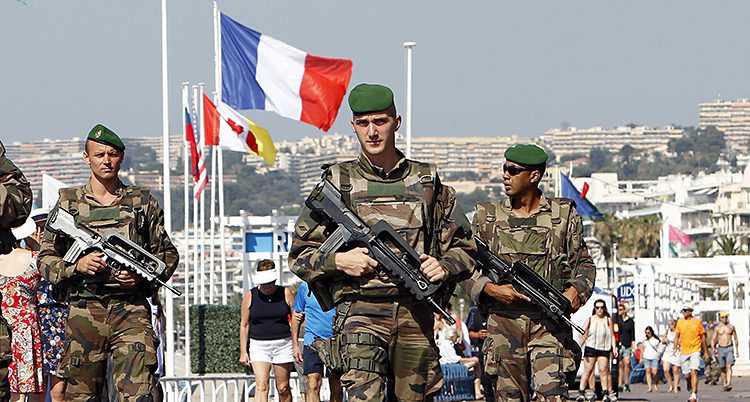 Tre soldater med vapen går efter gatan. I bakgrunden syns flaggor på halv stång.