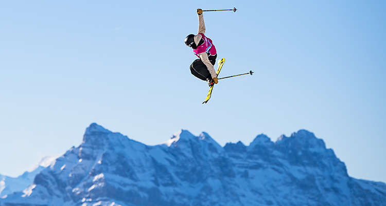 En person är högt uppe i luften med skidor. I bakgrunden syns stora fjäll med snö.