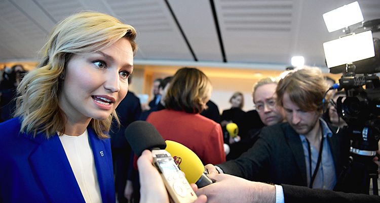 Kristdemokraternas ledare Ebba Busch blir intervjuad av flera journalister. Hon är i riksdagen. Hon pratar in i flera mikrofoner och det står folk med kameror runtomkring. Ebba Busch har blont hår och en blå kavaj.