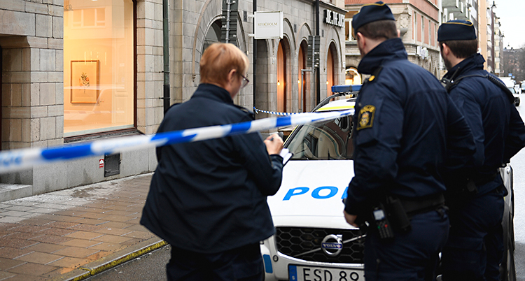 Bilden är tagen på en gata i Stockholm. Vi ser tre poliser med ryggen mot kameran. Vi ser också en polisbil och ett band från polisen som spärrar av mot ett hus. Bandet är blått och vitt.