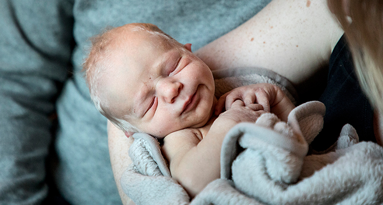 En nyfödd bebis ligger i en kvinnas famn. Bebisen har en grå filt runt sig.
