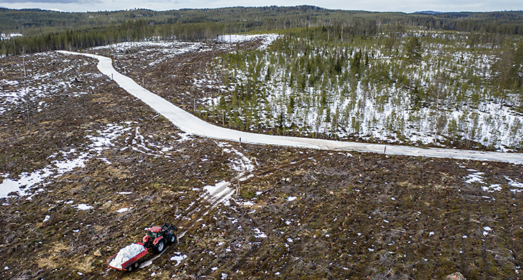 En flygbild som är tagen över skogen i Dalarna. En traktor kör snö i sin skopa. Traktorn kör mot ett spår för längdskidor.