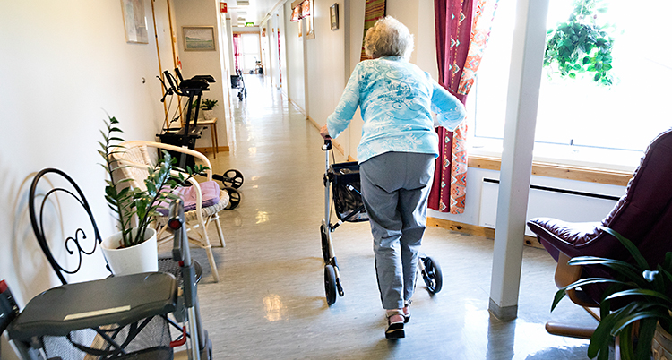 Bilden är tagen på ett boende för äldre människor. En kvinna går i en korridor med rullator. Hon har ryggen mot kameran.