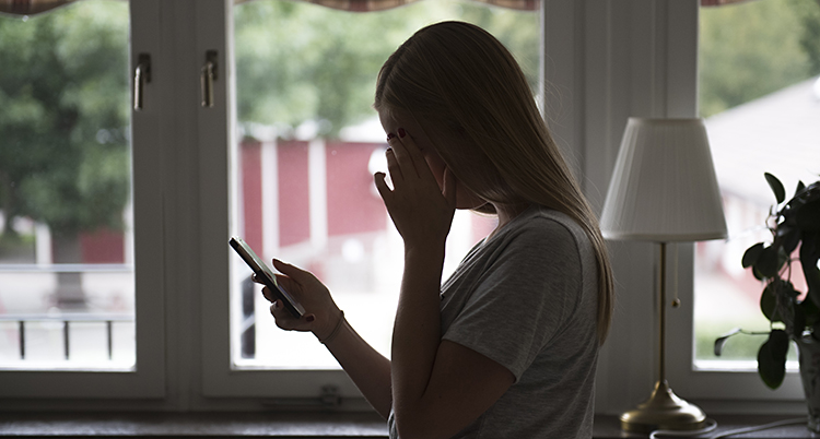 En ung tjej står inne i ett hus. Hon står framför ett fönster och tittar på sin mobiltelefon.