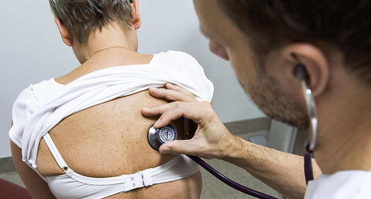 En kvinna sitter med uppdragen tröja med ryggen mot en läkare som lyssnar med ett stetoskop mot ryggen.