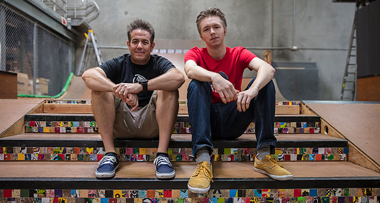 Två män sitter i en trappa. Mannen till vänster har svart t-shirt och grå shorts. Man till höger har röd t-shirt och blå jeans.