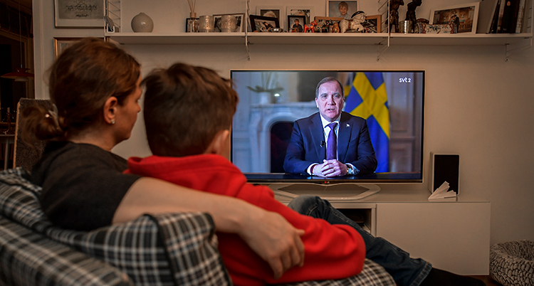 Ett vardagsrum i ett hem. En kvinna och en pojke sitter i en soffa. De tittar på tv.