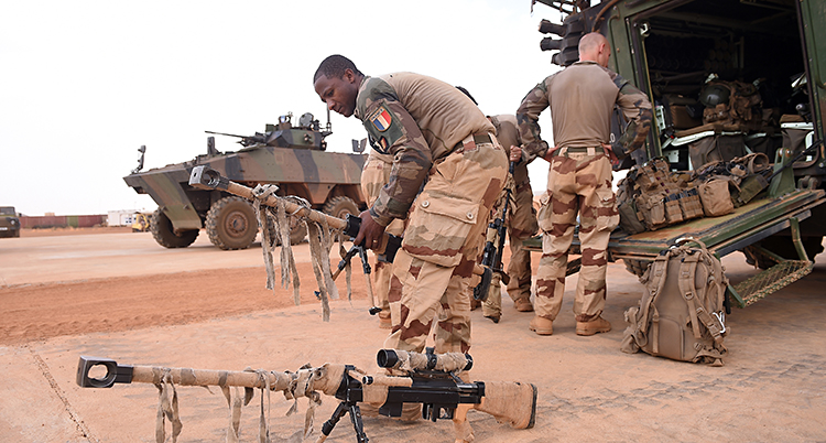 Några militärer i sandfärgade kläder tar fram stora vapen och ställer på marken. Bakom står en bandvagn. Marken är sandfärgad.