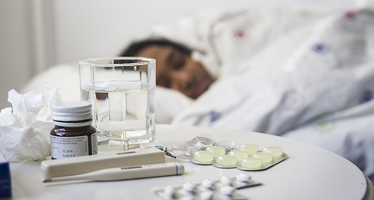 En kvinna ligger i sängen. Framför henne på ett bord finns febertermometer och tabletter mot värk.