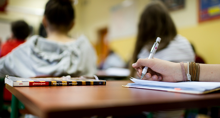 en elev skriver vid sitt bord i ett klassrum. Ryggarna av två andra elever syns.