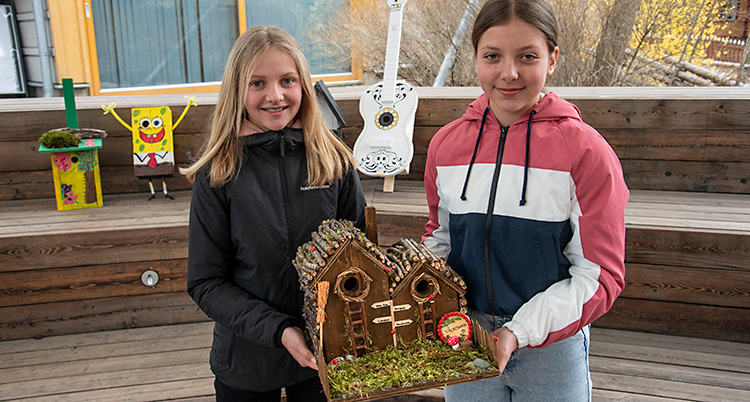 Två flickor visar en fågelholk som ser ut som ett hus av trä.