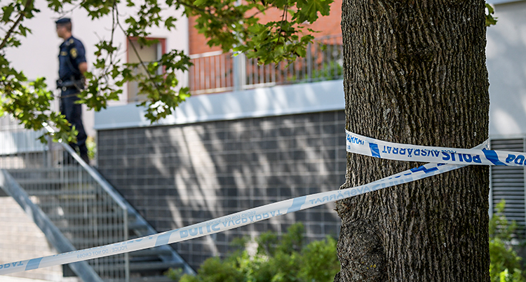 Bilden är tagen utomhus. Det är sommar. Man ser ett lägenhetshus. Poliserna har sprärrat av platsen med vita och blå band. I bakgrunden står en polis.