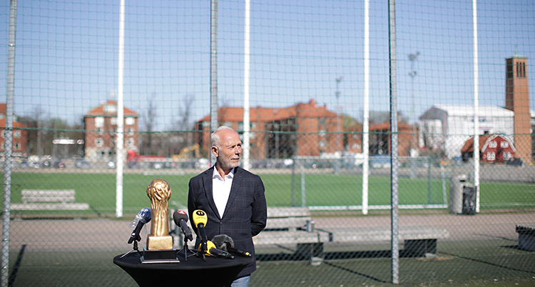 En man står framför en fotbollsplan. Han har flera mikrofoner framför sig på ett bord.