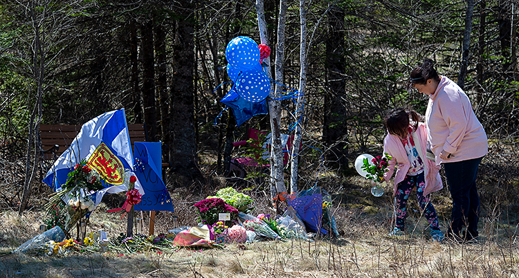 Bilden är tagen utomhus. En kvinna och ett barn står vid en plats. Där har folk lämnar blommor och gåvor för att hedra en dödad person.