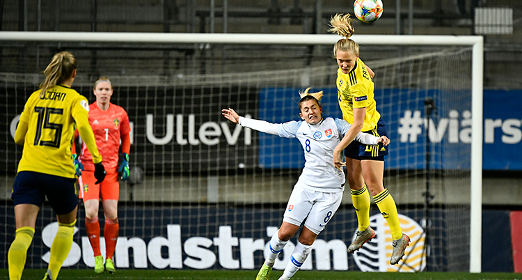 Bilden visar en match i fotboll. Två spelare hoppar för att nicka en boll. Den svenska spelaren hoppar högst.