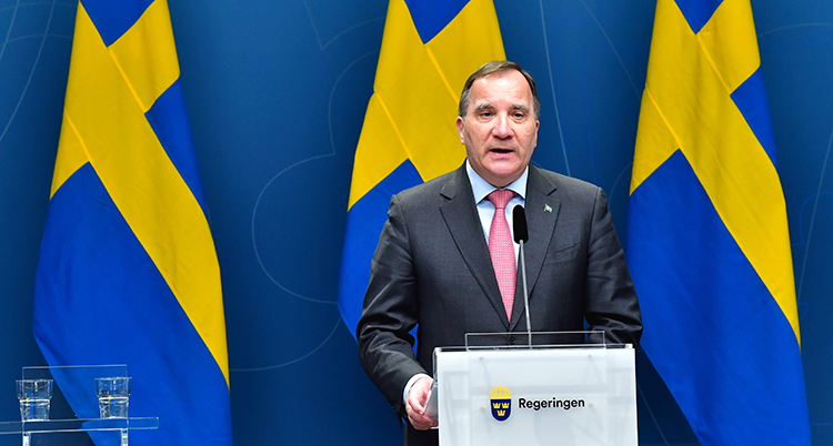 Han har en mikrofon framför sig. I bakgrunden syns Sveriges flagga.