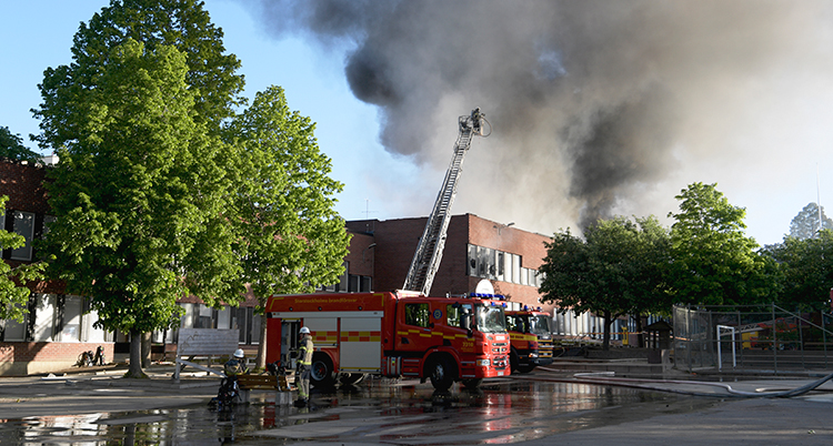 En bild på skolan från håll. Framför skolan står en brandbil med hissad stege över taket på skolan. Rök syns stiga upp mot himlen.