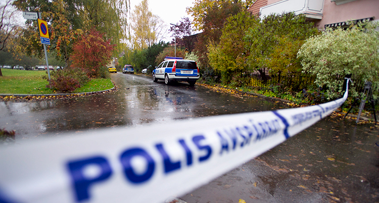 En polisbil står på en gata som är våt av regn. Det är buskar och låga hus. Längst fram på bilden är ett band där det står Polis. Det spärrar gatan.