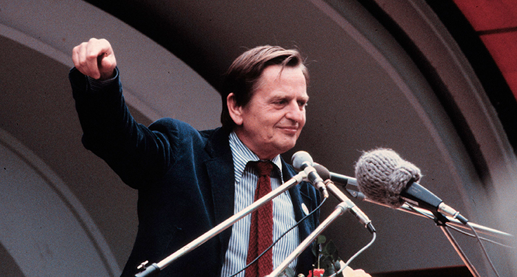 Olof Palme i en talarstol. Han ler lite och lyfter ena armen.