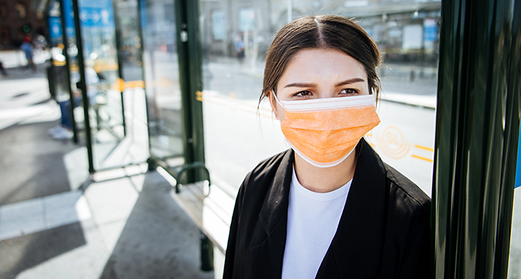 En ung kvinna står vid en busshållplats. Hon har på sig ett munskydd som är orange. Hon har en vit tröja och en svart jacka.