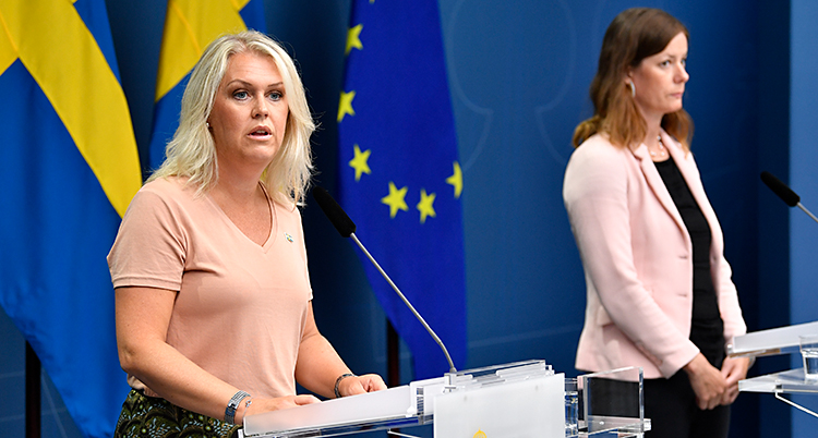 Två kvinnor har ett möte med journalister. De står vid varsitt bord och pratar. De har varsin mikrofon. I bakgrunden finns en svensk flagga och en EU-flagga.