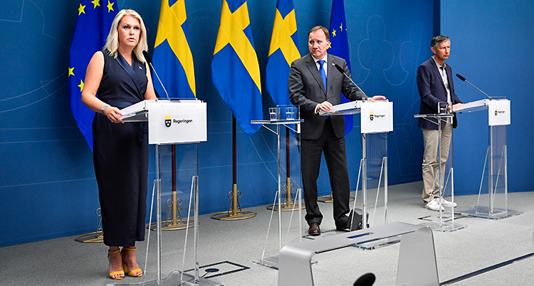Det är en träff med journalister. Längst till vänster står minister Lena Hallengren. I mitten står statsminister Stefan Löfven. Till höger står Richard Bergström. De har varsin mikrofon. I bakgrunden syns Sveriges flagga och EUs flagga.