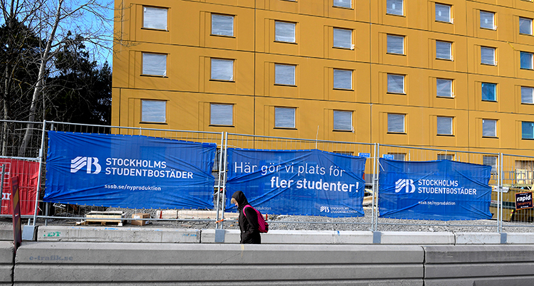 en person går utanför en studentbostad i gult. Framför bostaden står skyltar om att de bygger fler bostäder.