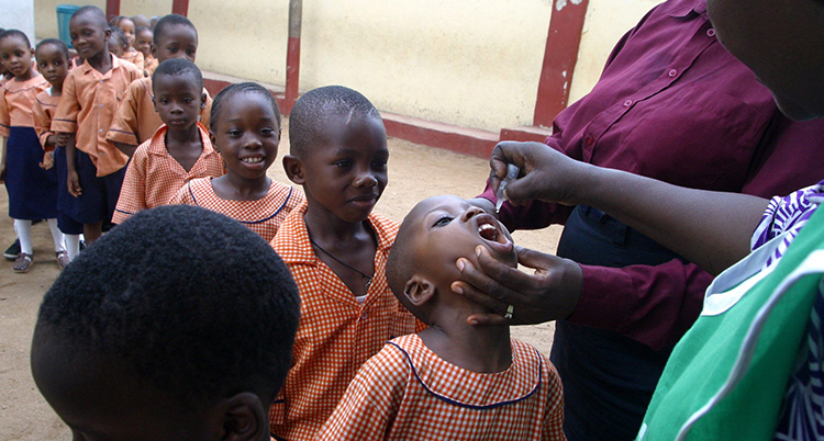 barn står på rad för att få vaccin. Barnet längst fram i kön får vaccin i munnen av en vuxen. Barnen är i sexårsåldern. De har alla samma röda skoluniform.