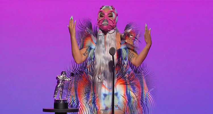 Lady Gaga står på en scen. Hon pratar i en mikrofon. Hon har en klänning som skimrar i olika färger. Och en orange gasmask för ansiktet.