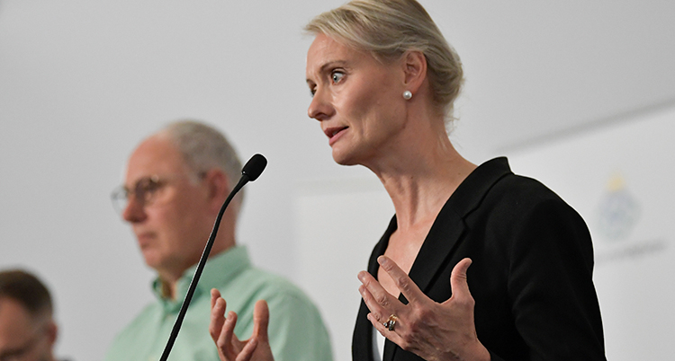 Karin syns i förgrunden på presskonferensen. Andra myndighetspersoner skymtas vid sidan om henne. Karin gör en gest med händerna.