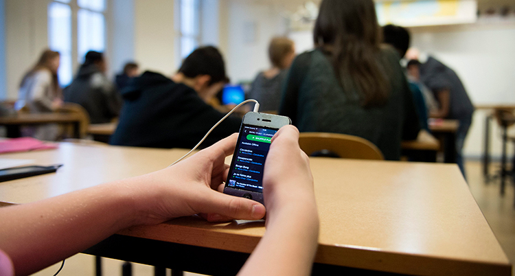 En elev som håller i en mobiltelefon. Men bara händerna syns. I Bakgrunden flera elever i ett klassrum.