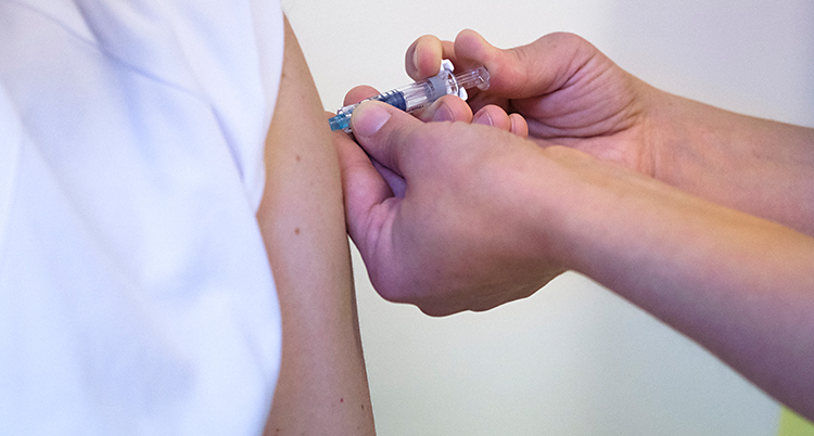 Vi ser hur en person får ett vaccin. En person trycker in en spruta i armen på personen som får vaccinet.