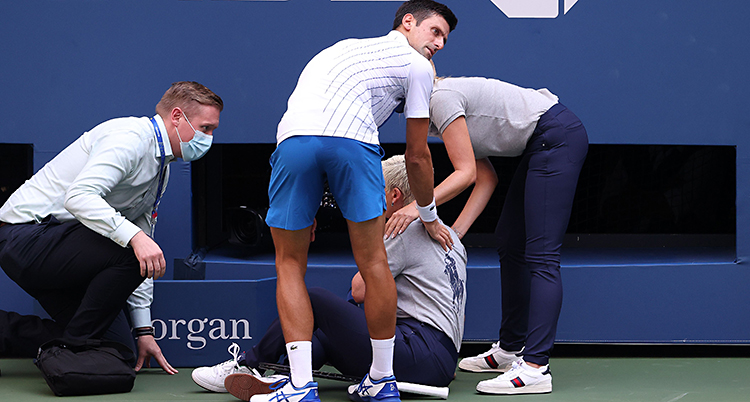 Vi ser en tennisplan. Novak Djokovic och två andra står runt en kvinna. Hon sitter på marken och tar sig mot halsen.