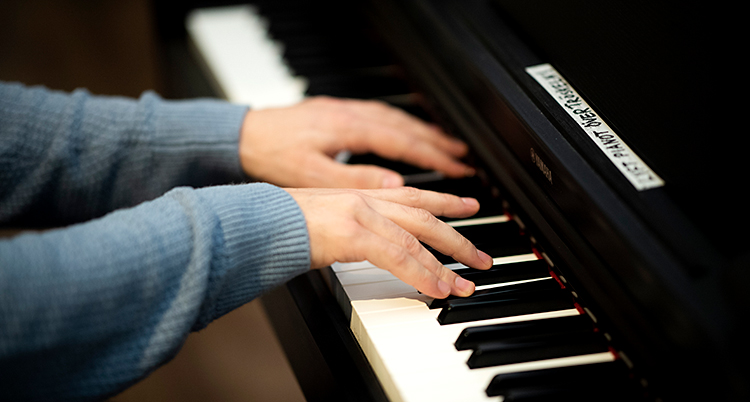 Vi ser armarna på en person som spelar piano. Personen har en grå tröja. Tangenterna på pianot är vita och svarta.